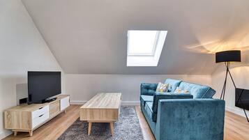 A modern loft apartment with a sloped ceiling, a skylight, a flat-screen TV, a wooden coffee table, a blue sofa, and a floor lamp.