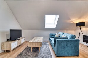 Un moderno apartamento tipo loft con techo inclinado, tragaluz, televisor de pantalla plana, mesa de centro de madera, sofá azul y lámpara de pie.