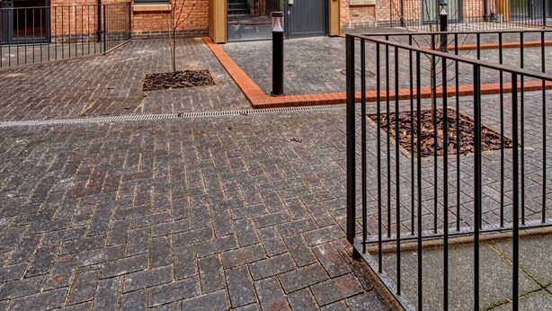 Un immeuble de plusieurs étages, construit en briques, avec une cour intérieure, comprenant une entrée centrale dotée de portes vitrées, une clôture en métal et un chemin piétonnier dallé.