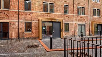A multi-story brick building with a courtyard, featuring a central entrance with glass doors, a metal fence, and a paved walkway.