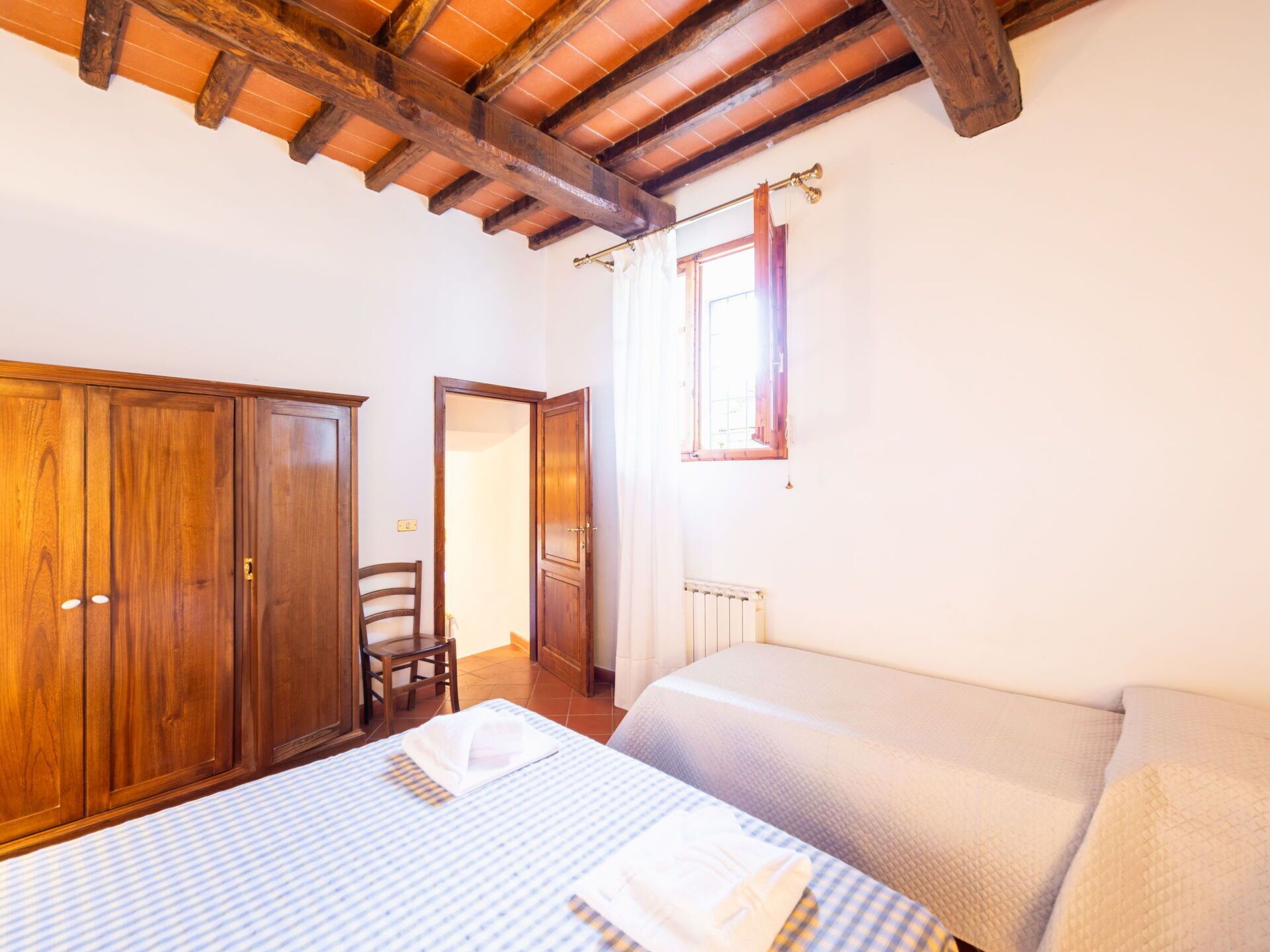 A bedroom with a wooden wardrobe, a chair, a bed with a quilted cover, and a window with curtains.