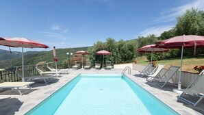 A rectangular pool with clear blue water, surrounded by white lounge chairs and red umbrellas, set against a backdrop of green hills and a clear sky.