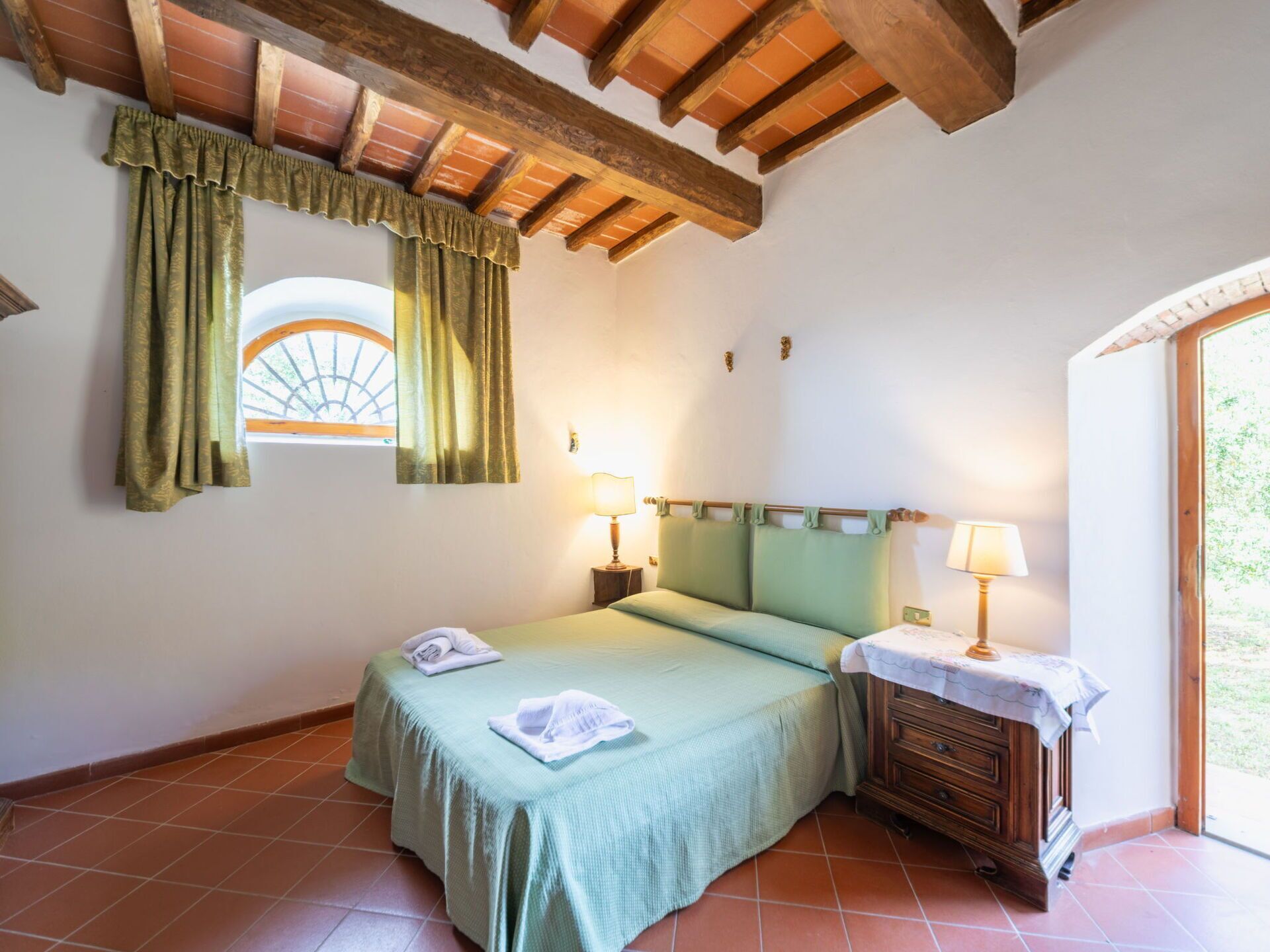 A bedroom with a bed, two bedside lamps, a wooden nightstand, a window with curtains, and a view of greenery outside.
