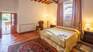 A bedroom with a bed, a chair, a nightstand, a window with curtains, and a radiator.