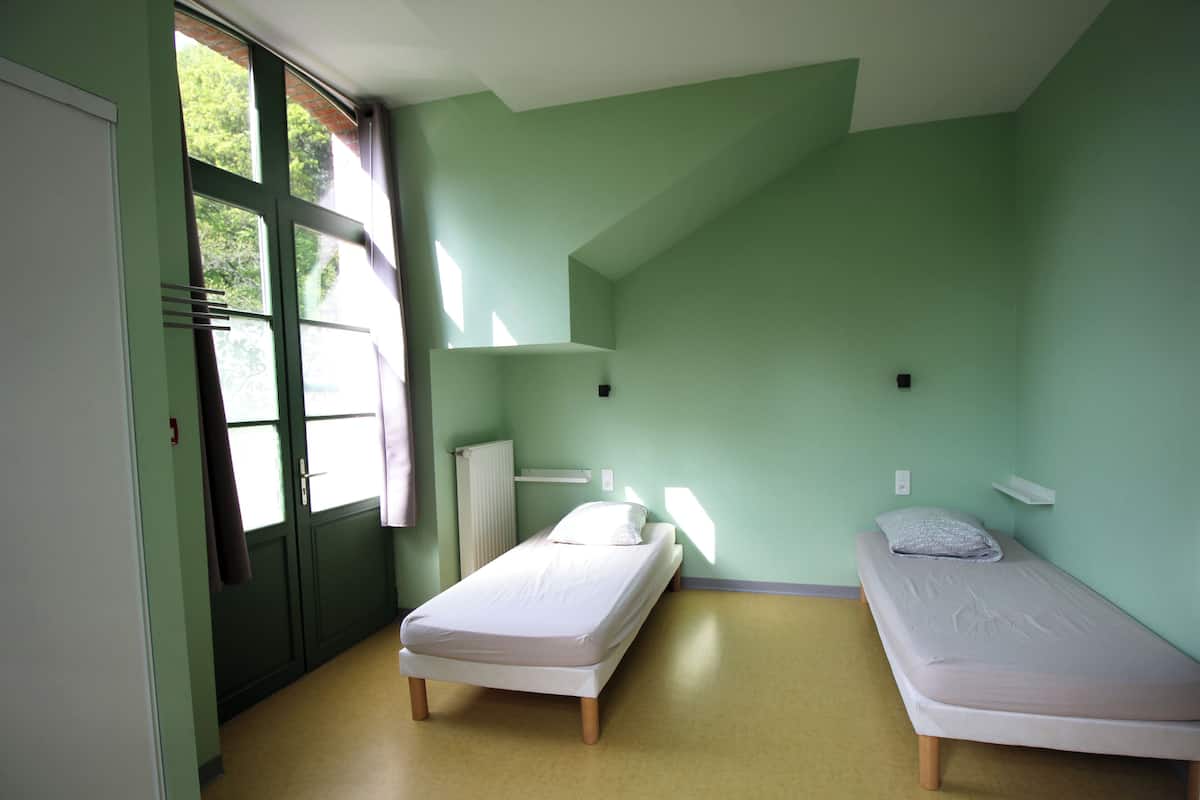 Una stanza con due letti singoli, un termosifone, una finestra con tende e una porta.