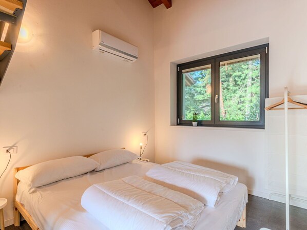 A bedroom with a bed, an air conditioner, a window with a view of trees, and a bedside table.