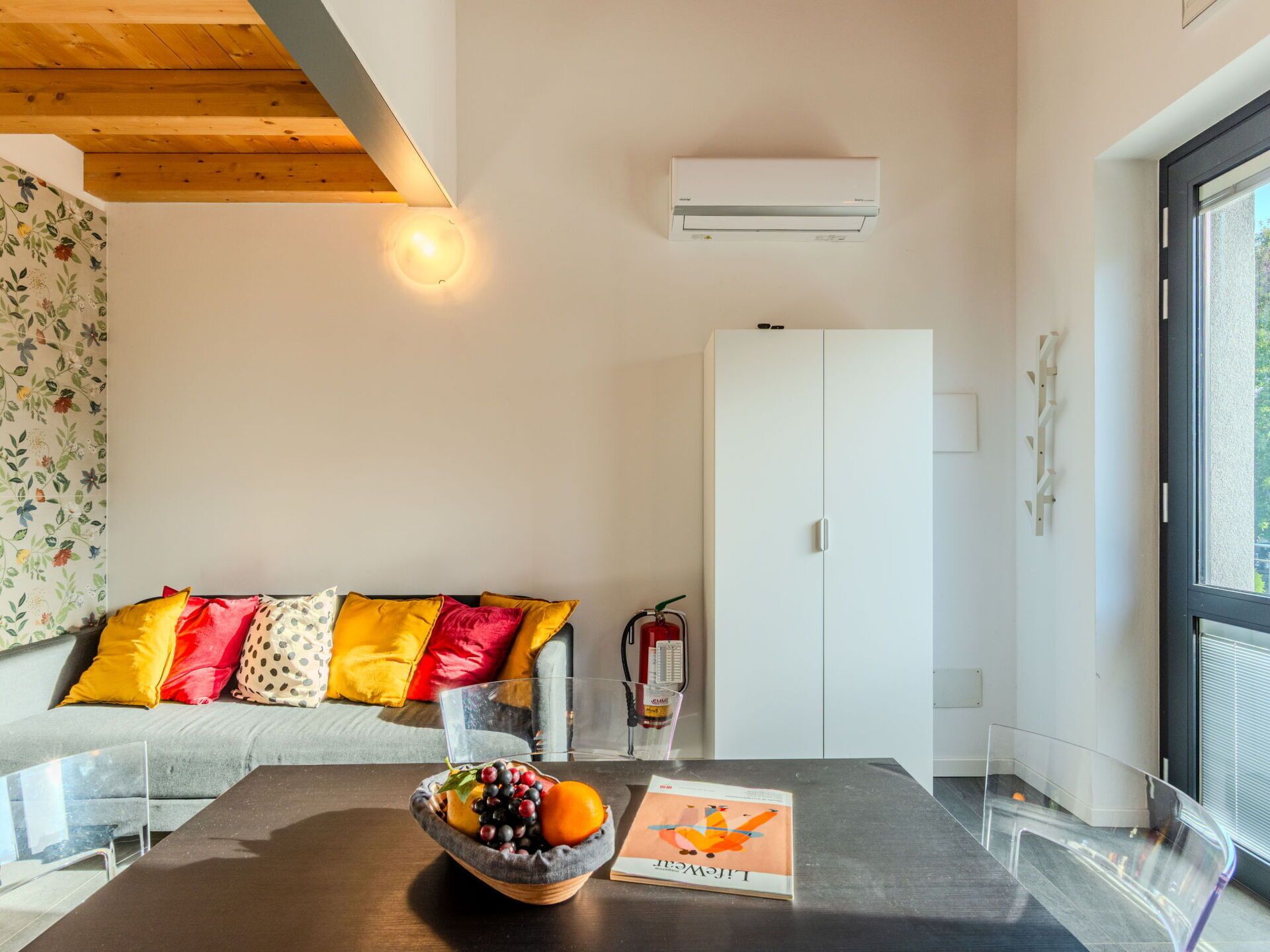 Een moderne woonkamer met een houten plafond, een witte kast, een glazen tafel met een fruitschaal en een tijdschrift, en bloemenbehang aan de linker muur.