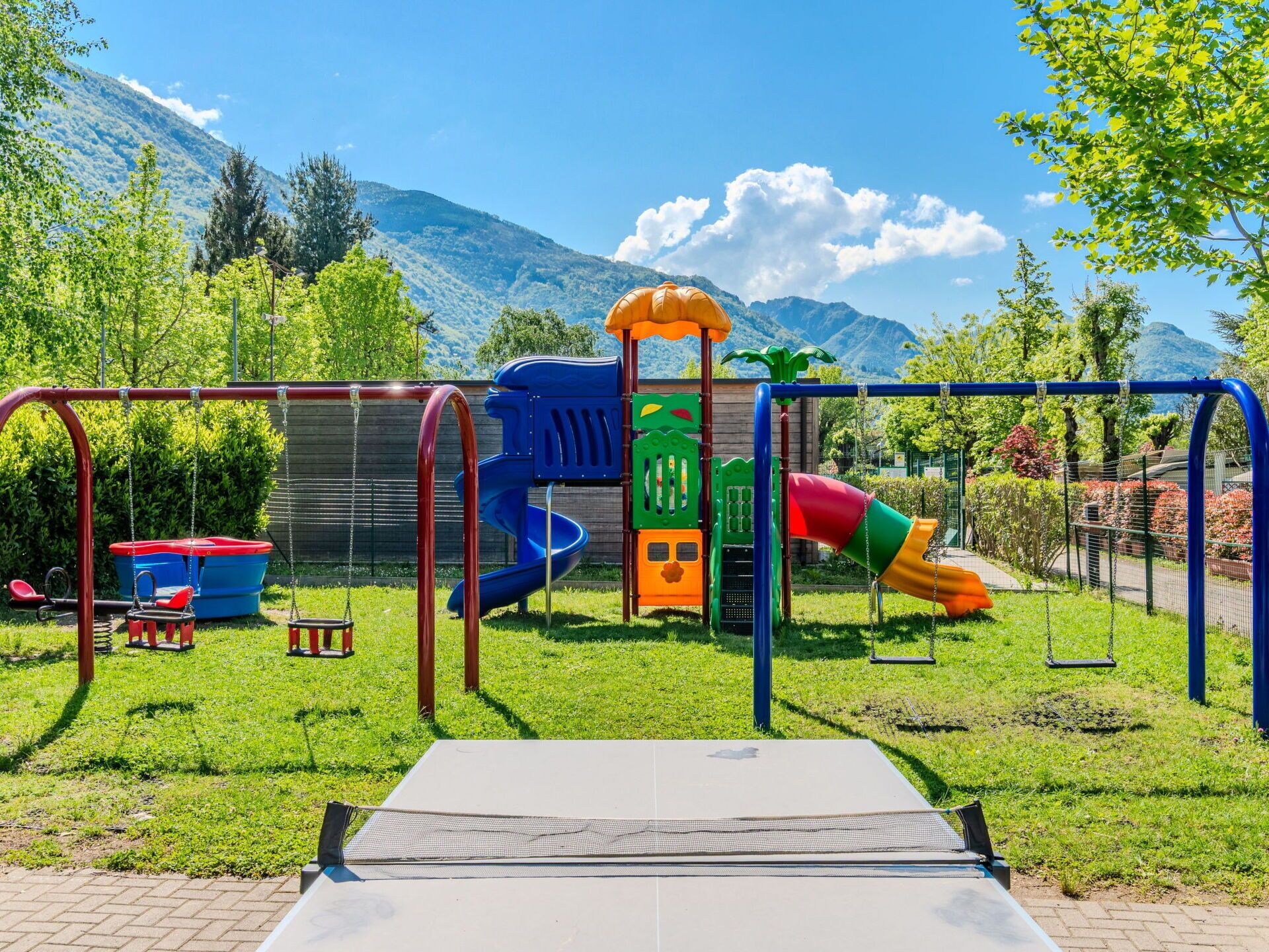 Een speelplaats met schommels, glijbanen en klimconstructies, omgeven door gras en bomen, met bergen op de achtergrond.