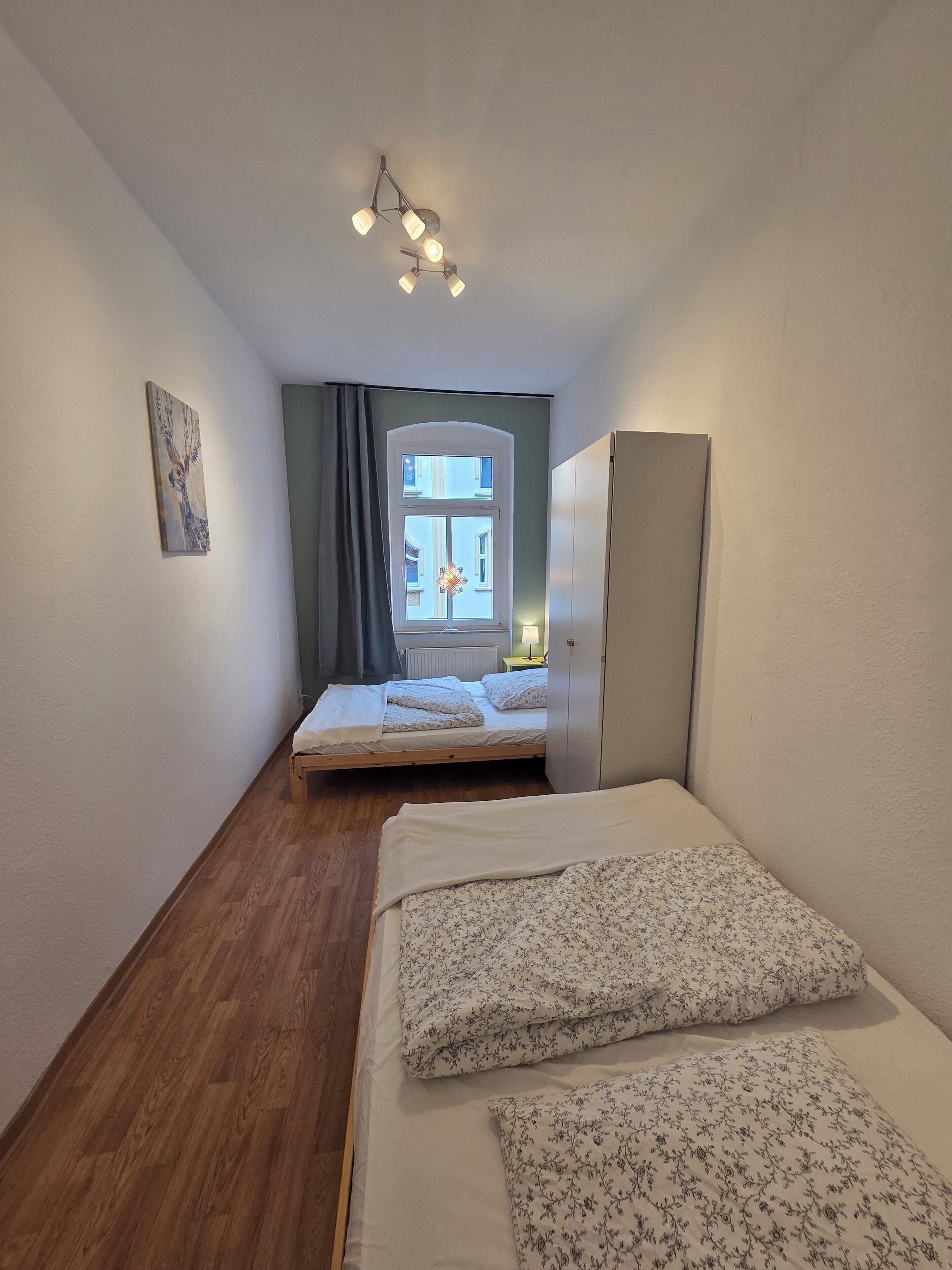 A narrow room with a bed, a wardrobe, a window with curtains, and a ceiling light fixture.