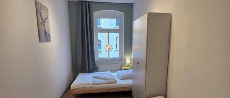 A narrow room with a bed, a wardrobe, a window with curtains, and a ceiling light fixture.