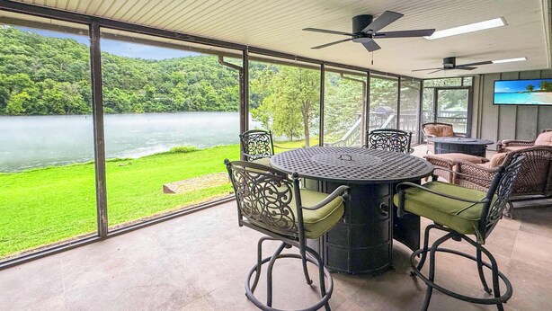 Ein Essbereich mit Tisch und Stühlen, großen Fenstern mit Blick auf einen See und Grünflächen, Deckenventilatoren und einem Flachbildfernseher an der Wand.