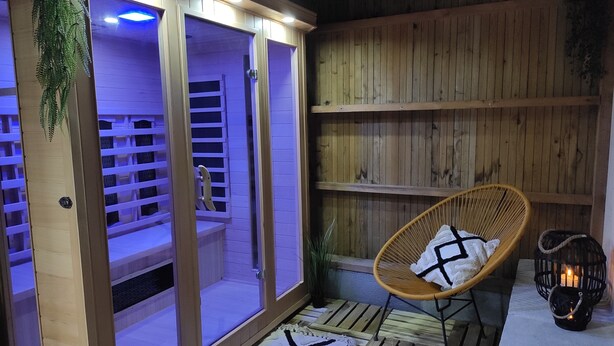 A wooden spa area with a glass-enclosed sauna, a wicker chair with a towel, and a lantern on the floor. - Gîte Le Karrdi (10 pers) in Brittany, ideal for families. Pets welcome! (Landavran)