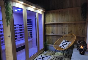 Uma área de spa em madeira com sauna envidraçada, uma cadeira de vime com toalha e um candeeiro no chão.