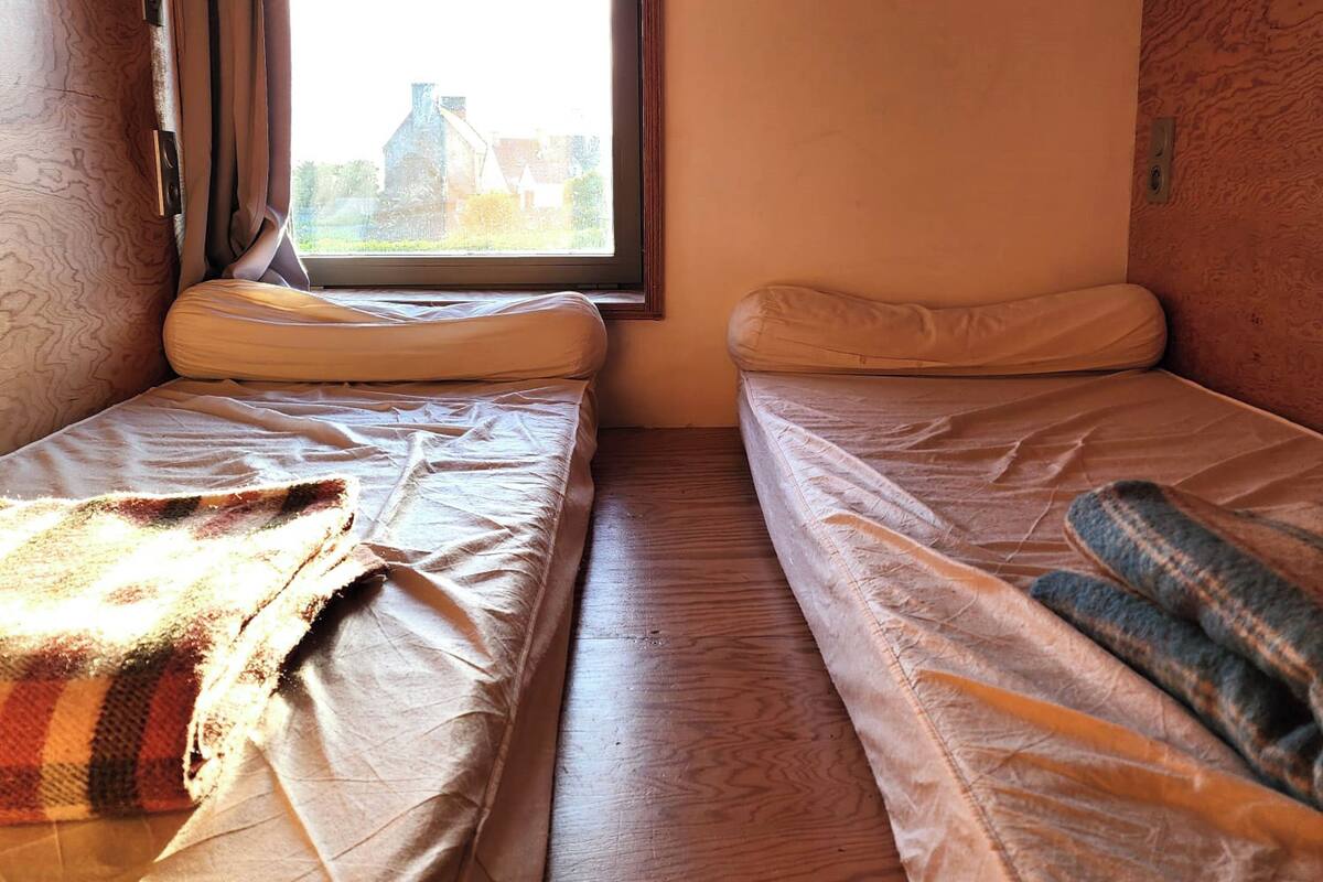A small, cozy room with two single beds, a window with a view of a building, and wooden walls.