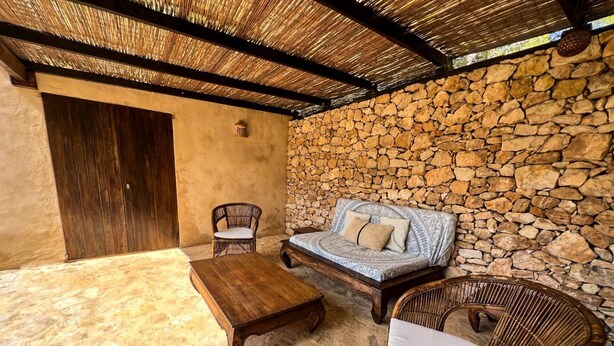 Una habitación con pared de piedra, puerta de madera, un banco tejido y sillas de mimbre.