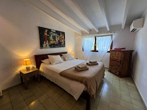 Un dormitorio con cama, mesita de noche, cómoda, un cuadro y una ventana con cortinas.
