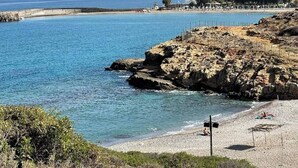 Una playa costera con terreno rocoso, aguas azules cristalinas y algunas sombrillas.