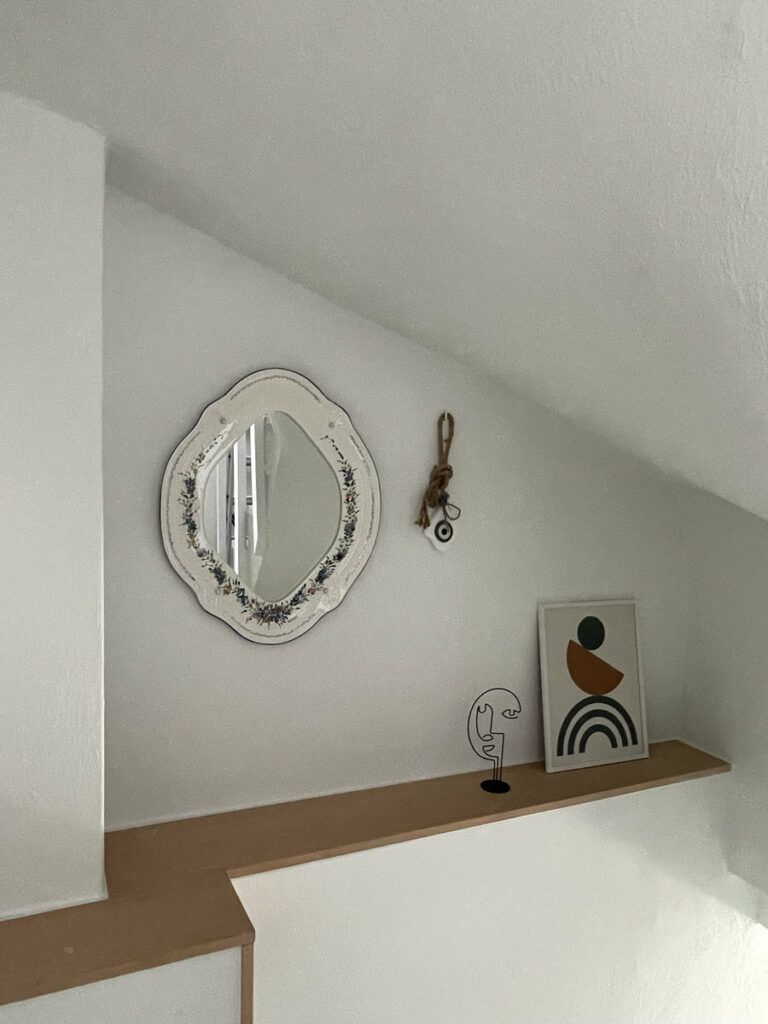Una pared blanca con un espejo redondo, un objeto decorativo pequeño y una obra de arte abstracta enmarcada sobre una repisa.