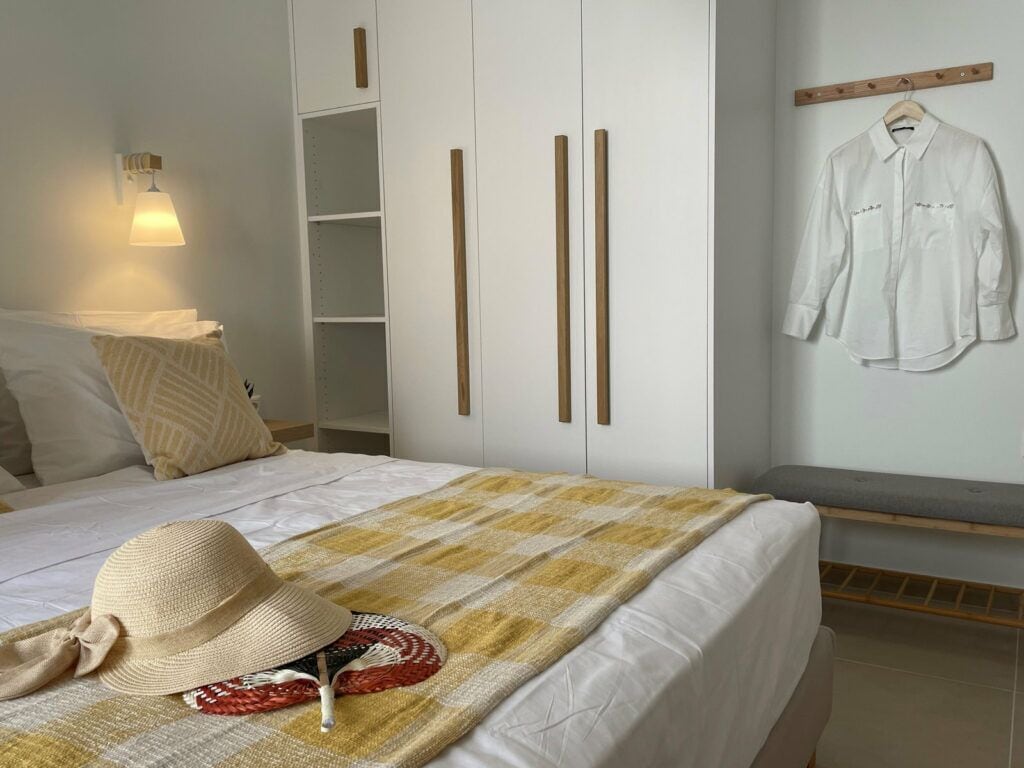 A neatly made bed with a checkered blanket, a hat, and a woven accessory on the bed. There is a white wardrobe with vertical handles and a hanging rack with a white shirt.