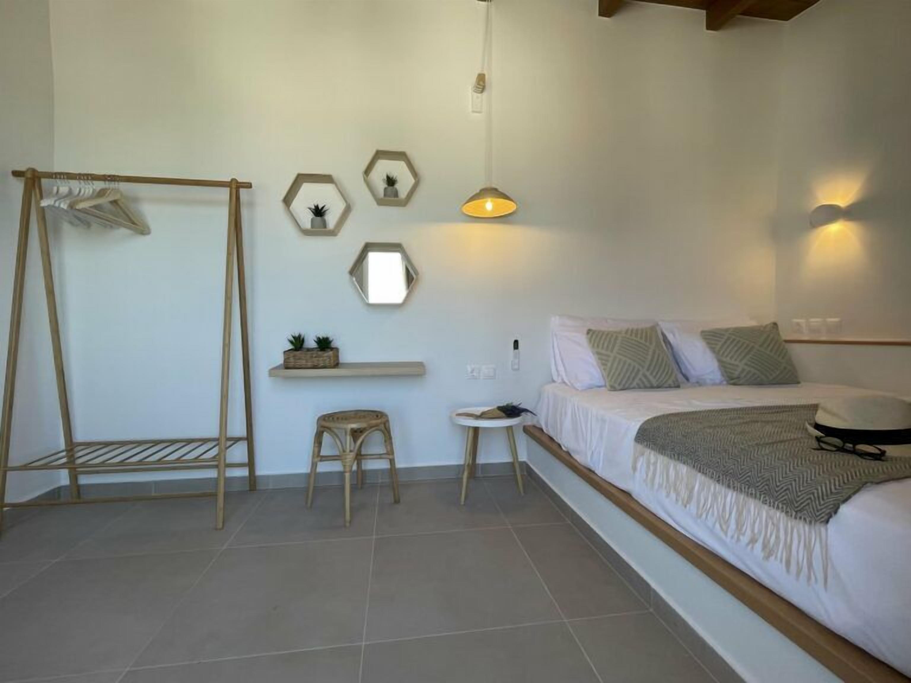 Un dormitorio con cama, una silla de madera, un taburete pequeño, una estantería con plantas y una lámpara de pared.