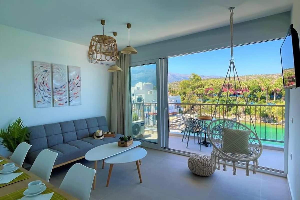 A modern living room with a grey sofa, a white coffee table, and wicker chairs. There is a balcony with a view of greenery and buildings.