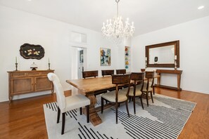 Une salle à manger avec une table en bois, des chaises, un lustre, un miroir et un tapis à motifs.