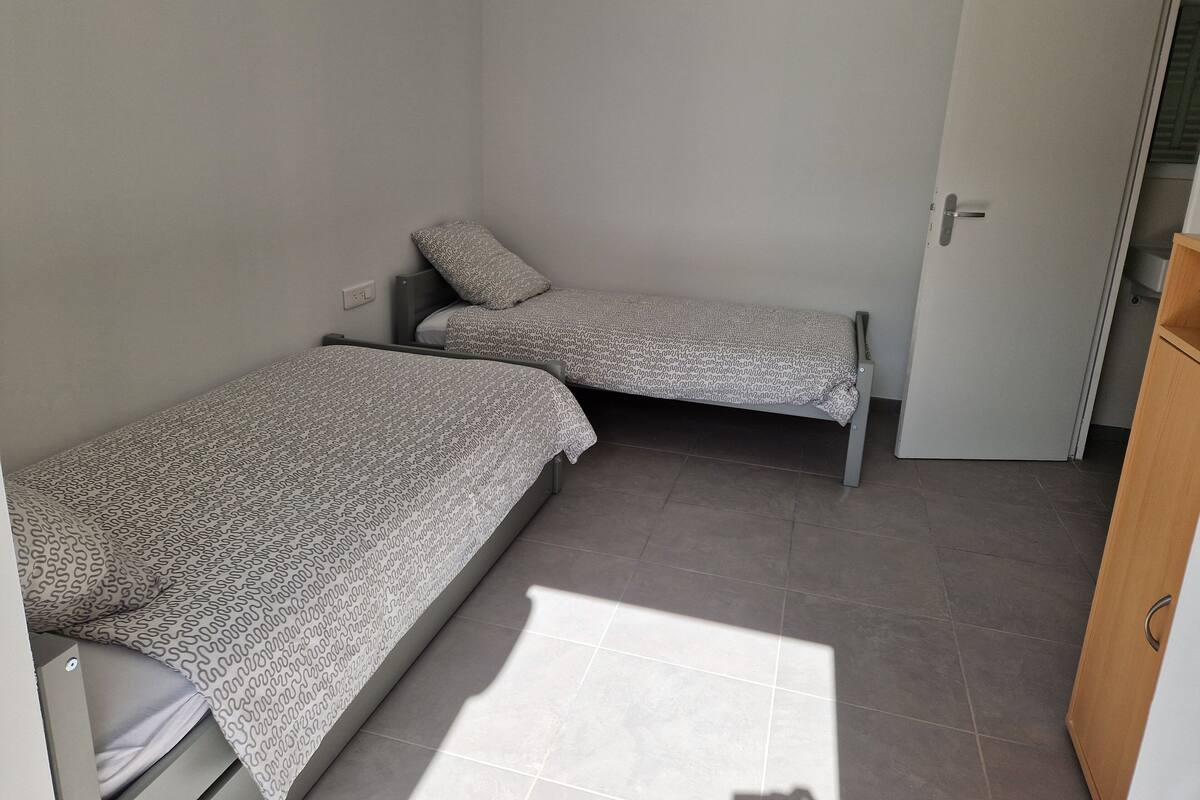Un petit logement composé d’une seule pièce, comprenant deux lits simples, une armoire en bois et un sol carrelé.