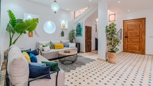A spacious living room with a sofa, coffee table, and potted plants. A staircase leads to an upper level, and a wooden door is visible. - Riad Dar Amarena - Exclusif - BreakFast (Marrakech)