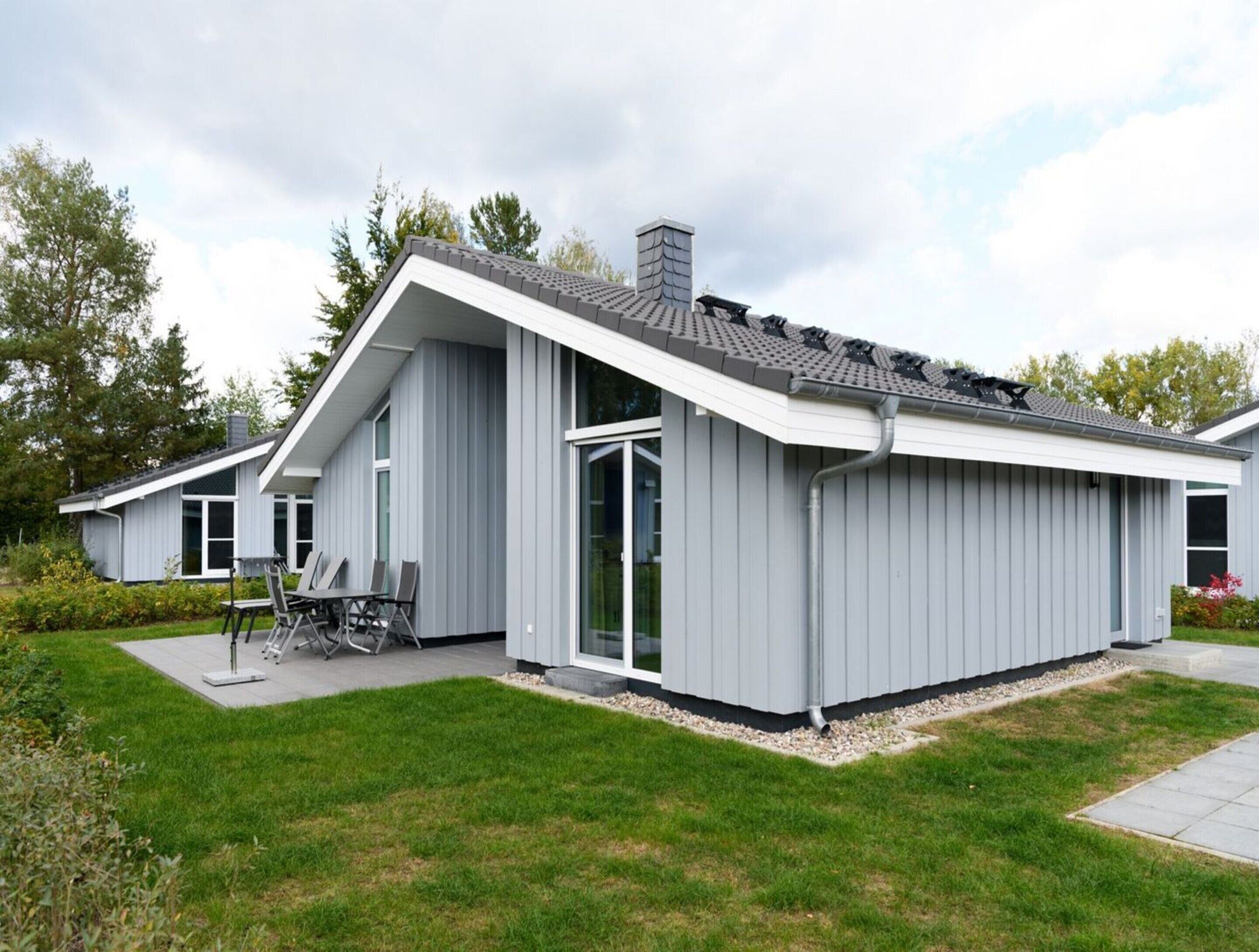 Ein modernes, zweistöckiges Haus mit geneigtem Dach, großen Fenstern und einer Terrasse mit Gartenmöbeln.