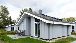 Ein modernes, zweistöckiges Haus mit geneigtem Dach, großen Fenstern und einer Terrasse mit Gartenmöbeln.