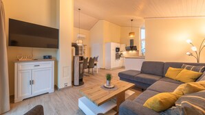 Ein modernes Wohnzimmer mit grauem Sofa, einem Holztisch und einem Essbereich mit einem weißen Schrank und einem Fernseher.