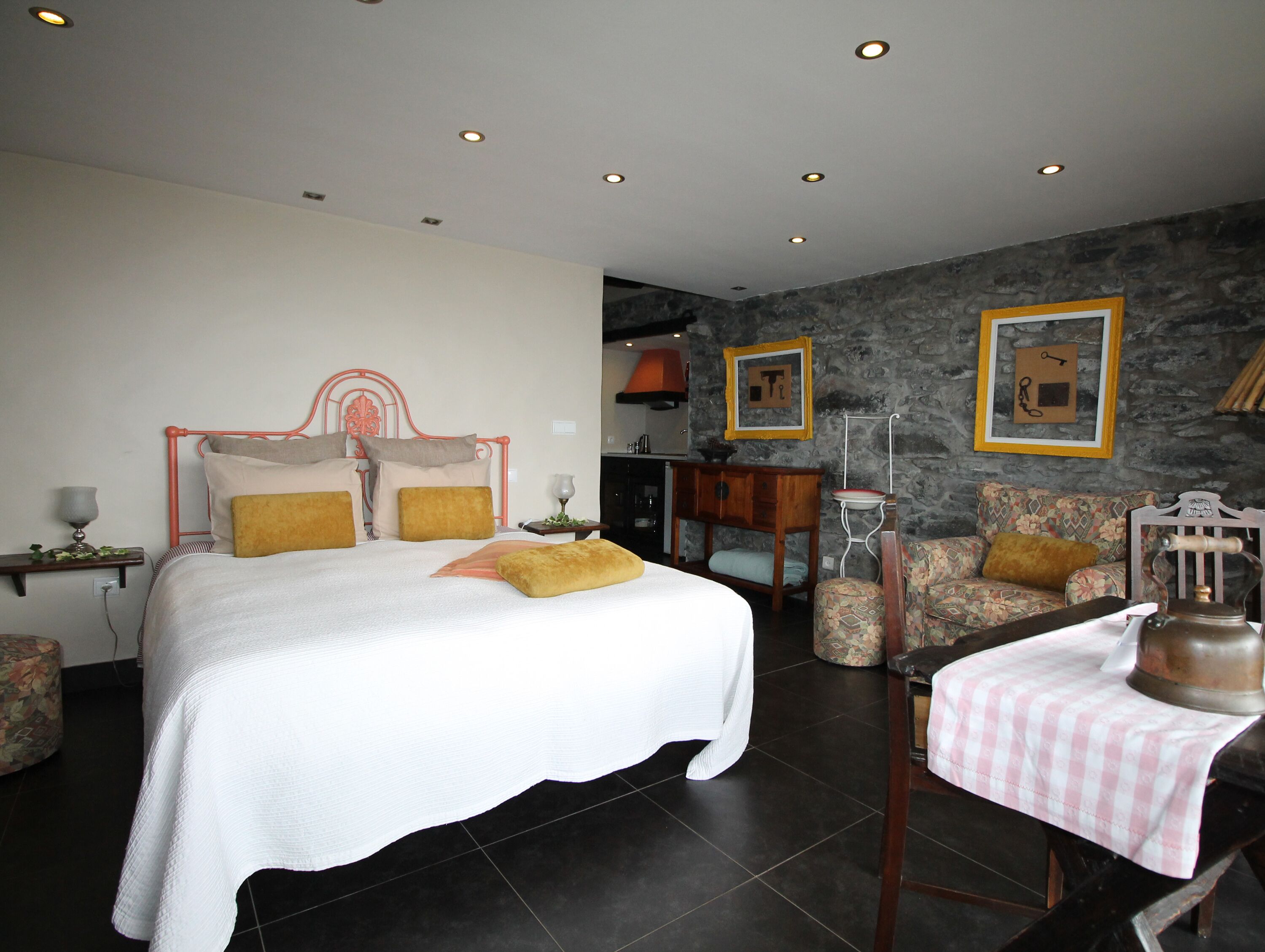 Quarto com cama, mesas de cabeceira, uma cadeira, uma mesa com chaleira e uma parede de pedra.
