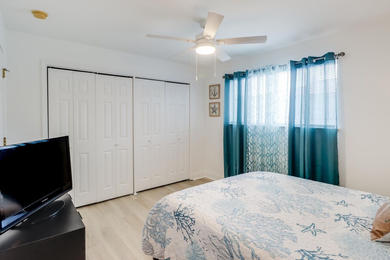A bedroom with a bed, a TV on a stand, a ceiling fan, and a wardrobe.
