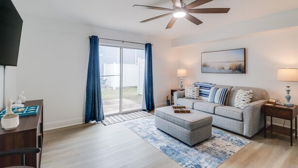 A modern living room with a grey sofa, blue curtains, a ceiling fan, and a large window.