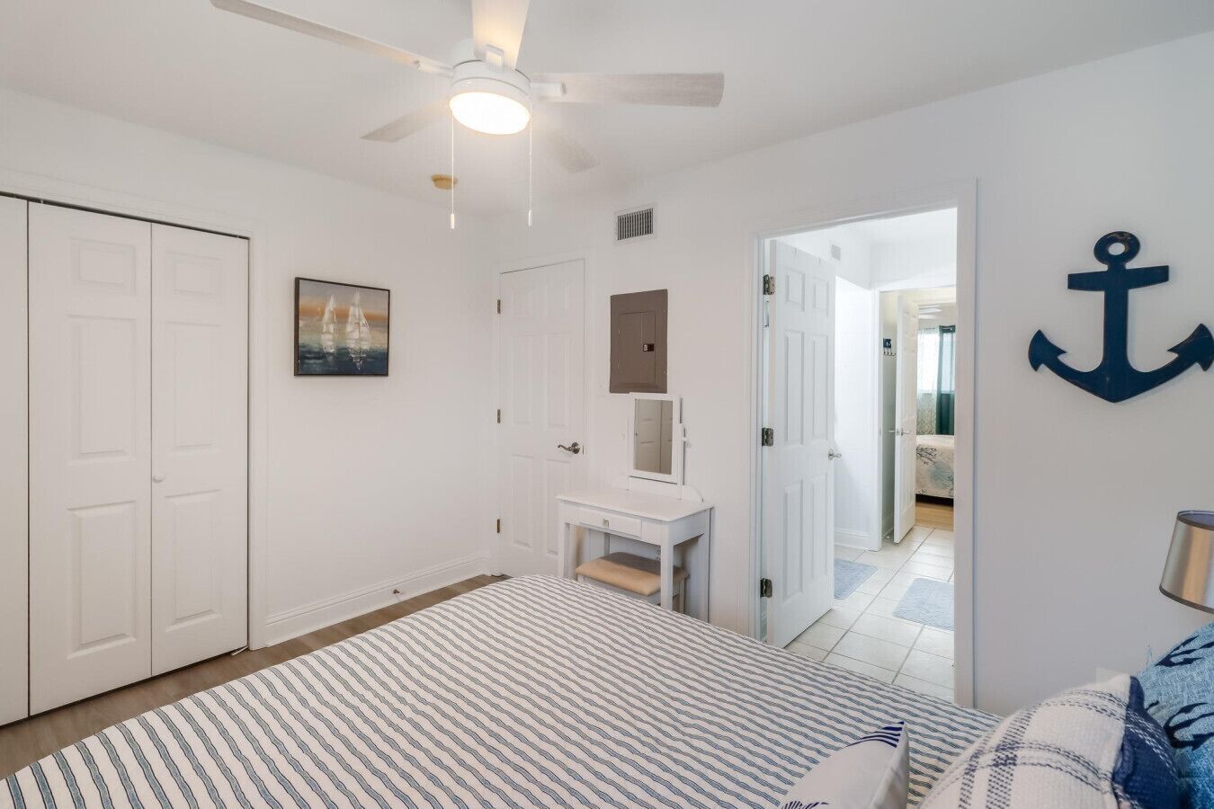 Un dormitorio con cama, mesita de noche, espejo, ventilador de techo y una puerta que da a otra habitación.