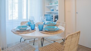 Un coin repas avec une table ronde dressée pour quatre personnes, entourée de chaises en osier. Une étagère blanche, sur laquelle on aperçoit de la vaisselle bleue et blanche, est visible à l’arrière-plan.