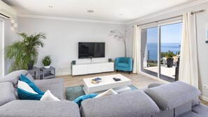 Una sala de estar moderna con un sofá gris, una mesa de centro blanca y un sillón azul. Un televisor de pantalla plana está montado en la pared, y hay un ventanal con vistas al mar.