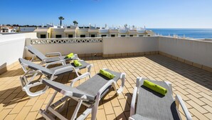 A rooftop terrace with lounge chairs and a red umbrella, overlooking a coastal area with buildings and the sea.