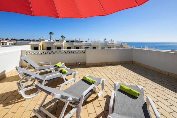 A rooftop terrace with lounge chairs and a red umbrella, overlooking a coastal area with buildings and the sea.