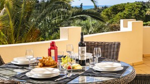 A table set for a meal with plates, glasses, and a bottle of wine, overlooking a scenic view of the ocean and palm trees.