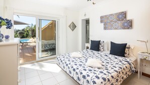 A bedroom with a bed, a nightstand, a bedside table, a window, and a view of a pool and garden.