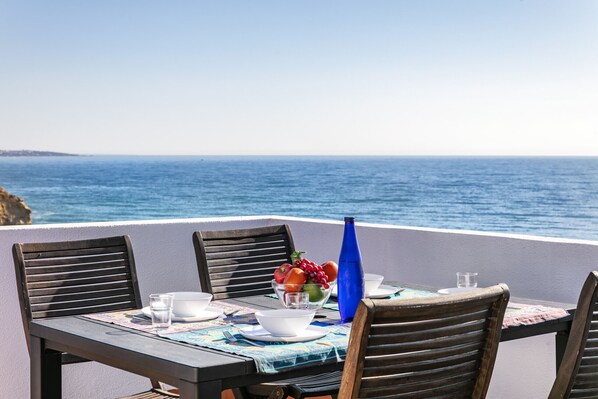 A table set with a bowl of fruit, a blue vase, and a glass of water, overlooking the sea.