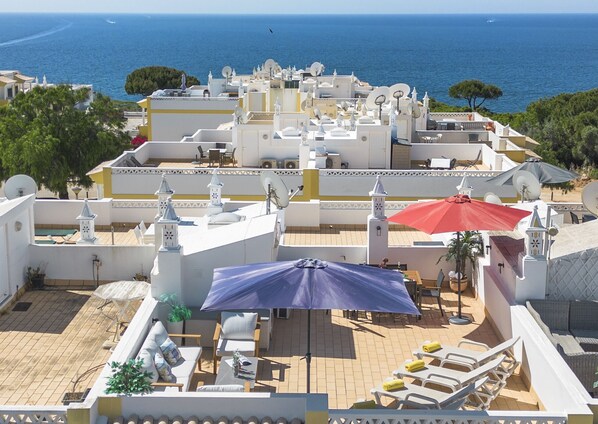 A rooftop terrace with white buildings, a pool, and a view of the sea.