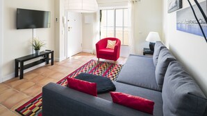 A modern living room with a grey sofa, red and black cushions, a red chair, a flat-screen TV, a black console table, a potted plant, and a colorful rug.