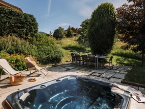 A hot tub with a table and chairs, surrounded by a garden and a hill in the background.