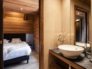 A modern bedroom with a wooden ceiling, a large bed, a nightstand, and a bathroom with a large sink and mirror.