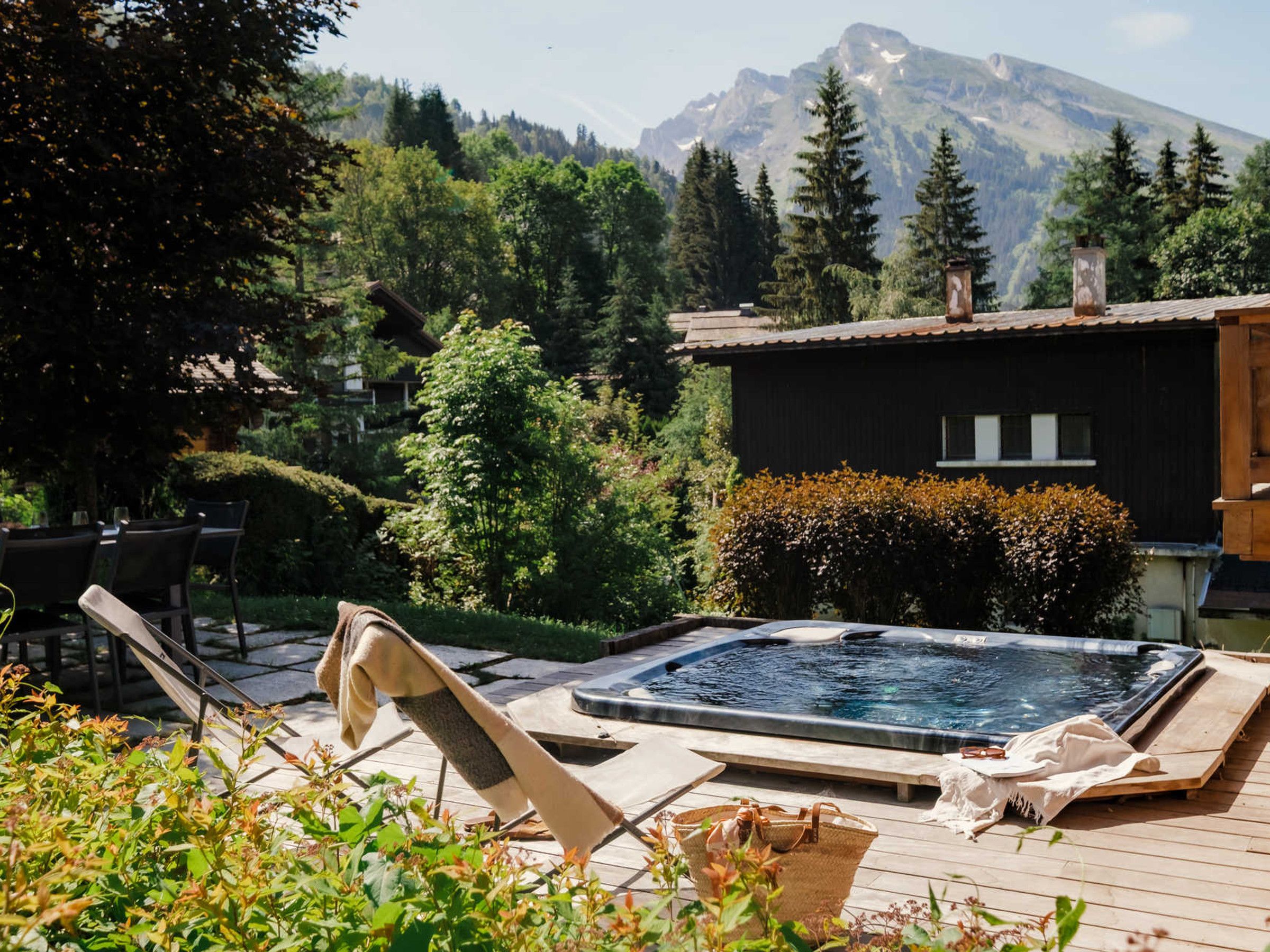 A hot tub in a garden with a mountain backdrop, wooden deck, and lounge chairs.