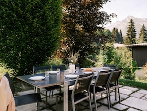 An outdoor dining area with a table set for four, surrounded by chairs, in a garden with trees and a mountain view.