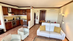 A living room with a white sofa and armchairs, a glass coffee table, and a kitchen area with wooden cabinets and modern appliances. - 06 Terraços de Benagil (Casa Rosa Azul) (Benagil)