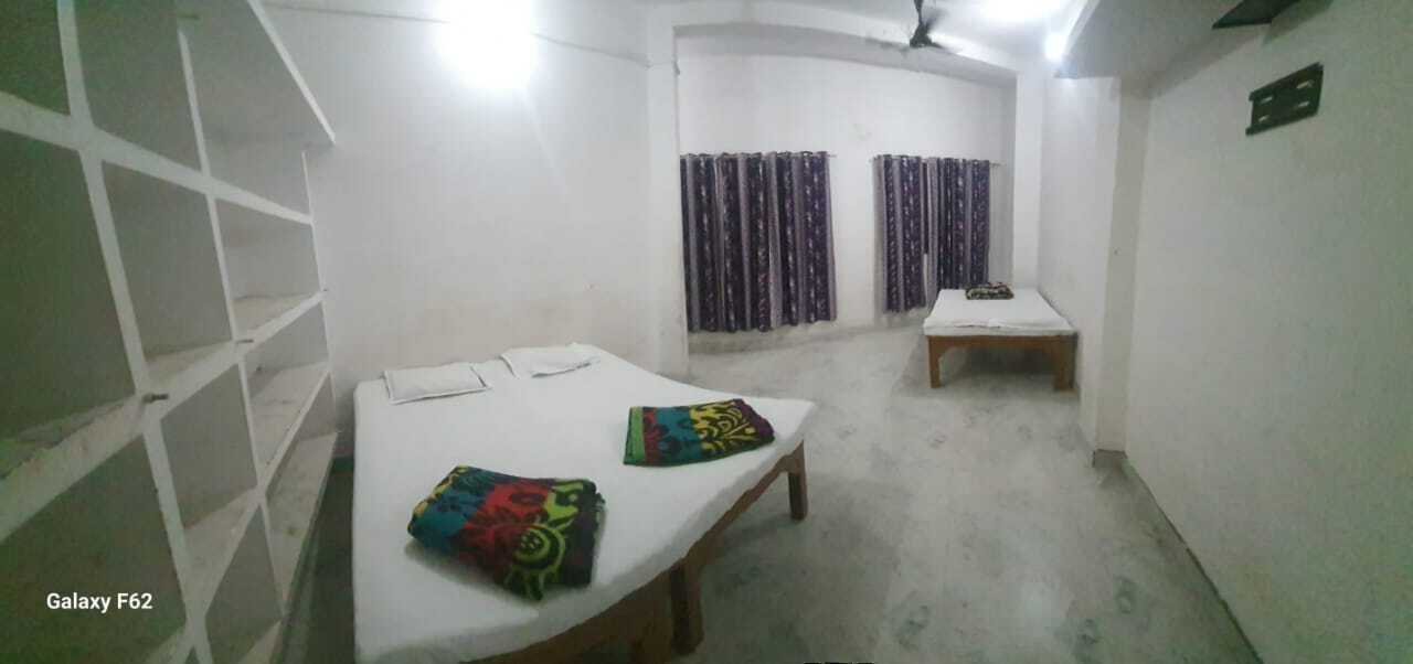 A small, dimly lit room with two single beds, a wooden bench, and a white wall with a shelf unit.